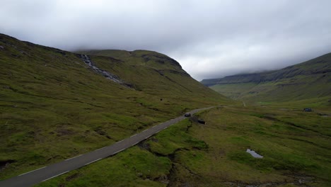 Car-parked-on-a-mountain-road-agaist-overcast-sky-in-Streymoy-next-to-a-cascade,-Faroe-Islands