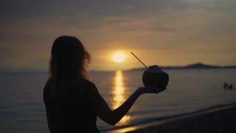 Movement-shot-of-a-women-holding-a-coconut-in-her-hand-at-a-beautiful-beach-at-sunset