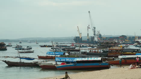 Barcos-Anclados-En-La-Playa-En-Stone-Town,-Zanzíbar,-Con-El-Puerto-De-Contenedores-Al-Fondo.