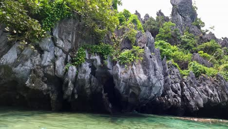 Tropical-island-tour-boats-moored-in-crystal-clear-turquoise-ocean-water-next-to-rugged-rocky-cliffs-in-El-Nido,-Palawan,-Philippines