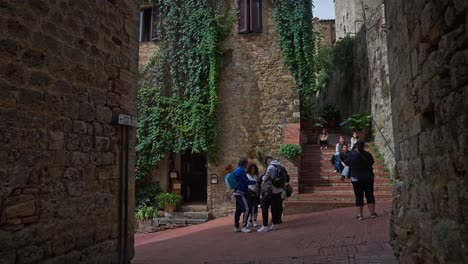 Gente-En-La-Calle-Con-Una-Casa-Cubierta-De-Plantas-De-Hiedra-En-La-Ciudad-Toscana-De-San-Gimignano,-Italia