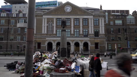 People-walk-past-a-huge-mound-of-rubbish-piled-up-around-a-statue-in-front-of-Tower-Hamlets-Town-Hall-as-a-refuse-truck-passes-by-following-two-weeks-of-refuse-strikes-in-the-borough