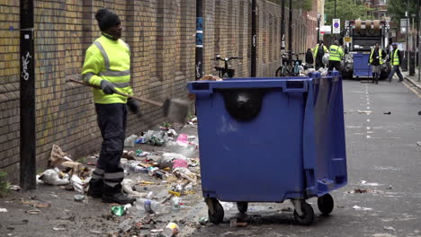 Un-Trabajador-De-Basura-Recoge-Basura-Con-Una-Pala-Y-La-Tira-En-Un-Contenedor-De-Basura-Azul,-Un-Hombre-En-Brick-Lane-Después-De-Dos-Semanas-De-Huelgas-De-Basura-En-El-Distrito-De-Tower-Hamlets.