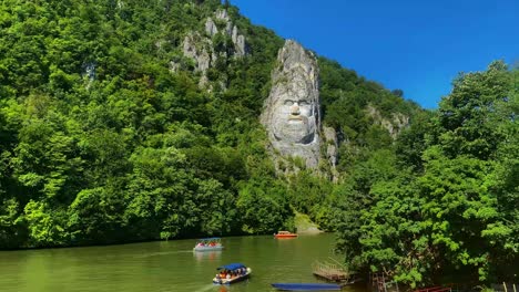 Boats-on-Danube-river-passing-around-colossal-carving-of-face-of-Decebalus-in-rock-above-water