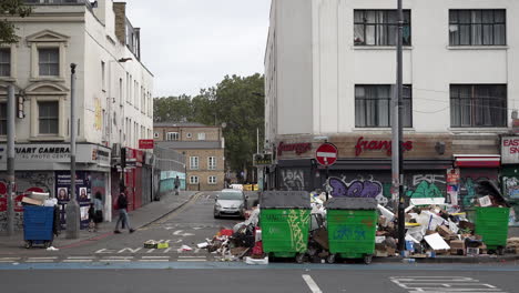 Nach-Zweiwöchigen-Müllstreiks-Im-Stadtteil-Tower-Hamlets-Laufen-Menschen-An-Müllhaufen-Vorbei,-Die-Sich-Um-Grüne-Mülltonnen-Auf-Der-Whitechapel-Road-Aufgetürmt-Haben