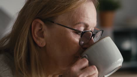 Extreme-close-up-of-thoughtful-caucasian-senior-woman-sitting-in-armchair,-drinking-coffee-and-looking-away