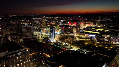 Aerial-approaching-shot-of-Atlanta-City-illuminated-at-night-with-high-rise-buildings,-ferris-wheel-and-Mercedes-Benz-Arena-in-background