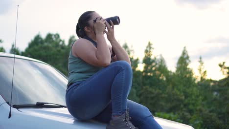 Woman-with-DSLR-camera-in-her-hands,-sitting-on-the-hood-of-her-car,-taking-photographs-outdoors-during-sunset