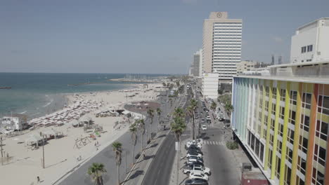 Push-in-drone-shot-to-the-side-of-The-colorful-Dan-Hotel-in-Tel-Aviv,-Rainbow-building-on-the-Gordon-promenade-and-Frishman-beach-full-of-visitors-on-a-warm-and-calm-summer-day