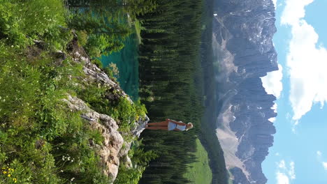 Beautiful-blonde-girl-standing-on-the-edge-of-cliff-with-a-panorama-view-of-Lake-Carezza-and-Dolomite-Mountains-in-Italy