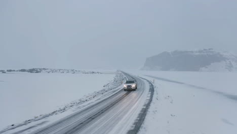 Aerial-view-backwards-in-front-of-a-SUV-driving-on-a-snowy-road,-blizzard-in-Iceland