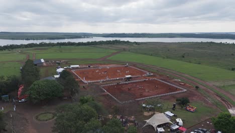 Vista-Aérea-Del-Emocionante-Evento-De-Salto-De-Caballo-En-El-Granero-Ubicado-Ajustado-A-Un-Cuerpo-De-Agua