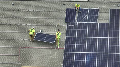 Workers-in-neon-shirts-and-harnesses-carrying-solar-panel-up-shingle-roof