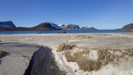 Aerial-dolly-forward-shot-skimming-over-the-sand-dunes-at-famous-Haukland-beach-in-Lofoten-Norway-in-late-winter-with-small-patches-of-snow-and-stunning-mountain-views-over-the-water