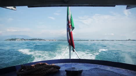 Rearview-of-boat-wake-driving-across-Lake-Garda-Italy-on-blue-sky-day,-Italian-flag-behind