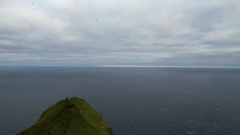 Aerial-shot-of-adventurous-hikers-stand-on-a-cliff-edge-taking-pictures-by-Kallur-lighthouse-in-Kalsoy,-Faroe-Islands