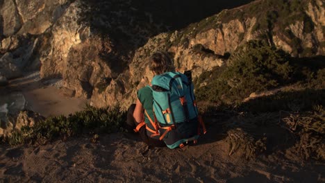 Back-view-of-female-tourist-with-backpack-sitting-at-cliff