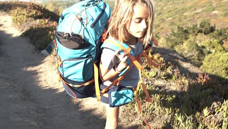 Tired-girl-walking-on-mountain-path,-carrying-big-heavy-backpack