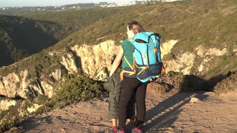 Mom-with-camping-backpacks-and-daughter-kid-standing-at-cliff