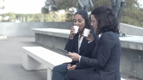 Businesswomen-sitting-on-bench,-drinking-coffee-and-talking