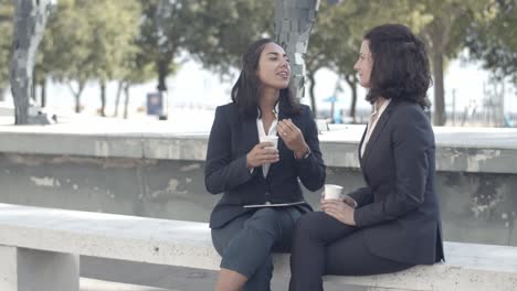 Two-businesswomen-sitting-outside,-drinking-coffee-and-talking