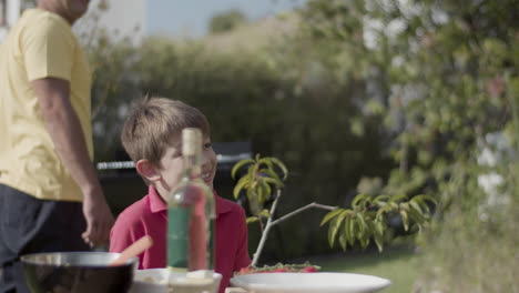 Smiling-preteen-boy-sitting-at-table-in-garden-and-smiling