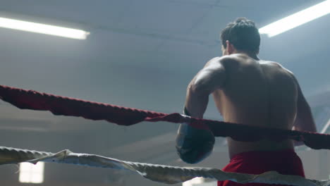 Junger-Männlicher-Boxer,-Der-Sich-Auf-Ein-Ringseil-Stützt-Und-Dann-Einen-Schattenkampf-Führt
