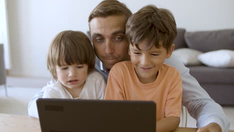 Happy-middle-aged-dad-hugging-sons-and-chatting-online-on-laptop