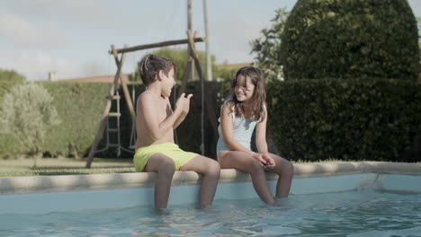 Caucasian-boy-and-girl-sitting-on-poolside,-talking-and-laughing