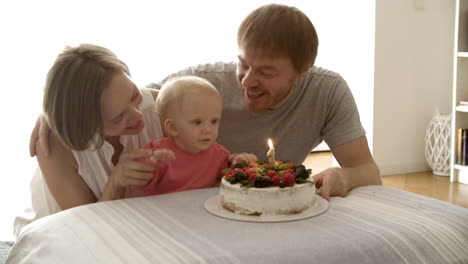 Fröhliche-Mutter-Und-Vater-Blasen-Mit-Ihrem-Kleinen-Mädchen-Kerze-Auf-Den-Kuchen