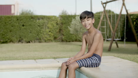 Asian-boy-sitting-on-pool-edge-smiling,-dangling-feet-in-water.