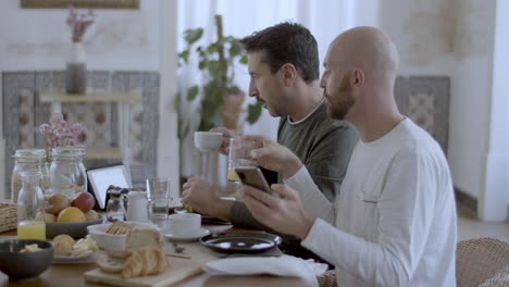 Gay-couple-using-gadgets-while-having-breakfast-in-kitchen