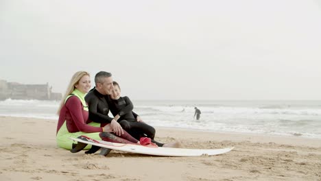 Glückliche-Familie-Mit-Surfbrett-Am-Strand-Sitzend
