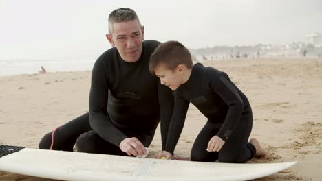 Happy-father-and-son-waxing-surfboard-on-beach