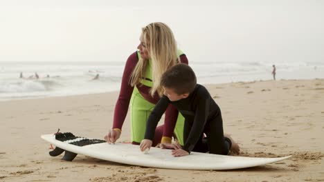 Happy-mother-and-son-waxing-surfboard-on-beach