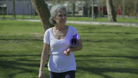 Front-view-of-woman-walking-in-park,-holding-yoga-mat-in-hand
