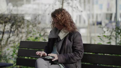 Concentrated-curly-woman-typing-on-tablet-while-sitting-on-wooden-bench