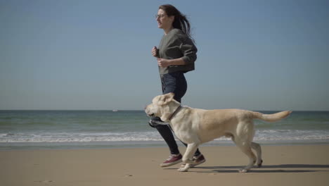 Happy-young-woman-in-eyeglasses-running-with-dog-on-seashore.