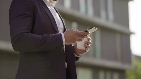 Businessman-holding-paper-cup-and-using-smartphone-on-street