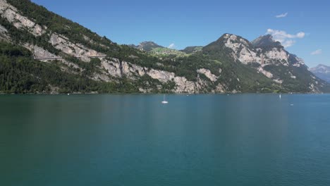 Wunderschöne-Landschaft-Am-Seeufer-Im-Sommer-In-Der-Schweiz,-Landschaftlich-Reizvolle-Natur-In-Den-Alpen,-Berge,-Eine-Yacht,-Ein-Weißes,-Strahlendes-Boot,-Das-Auf-Dem-Wasser-Schwimmt,-Und-Grüne-Walddecke-Auf-Felsigen-Klippenhügeln