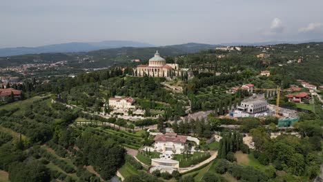 aerial-view-of-a-mountain-village-shows-a-large-building-with-a-domed-roof-and-circular-structure-on-top-of-the-hill-surrounded-by-lush-green-trees-more-buildings-and-mountains-stretch-out-horizon