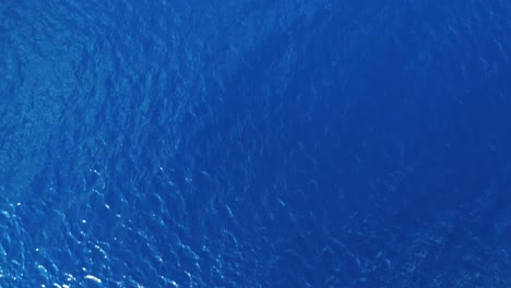 Drone-slowly-lowers-to-deep-blue-ocean-water-view-from-surface-as-ripples-and-waves-blow-across-top,-ocean-texture