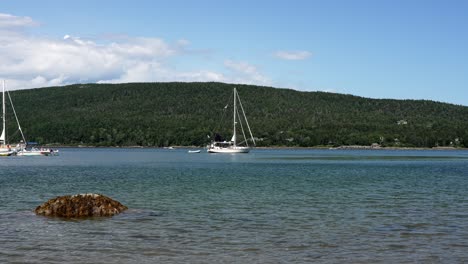 Sailboat-anchored-in-front-of-forested-mountainside-under-blue-sky-and-white-fluffy-cloud