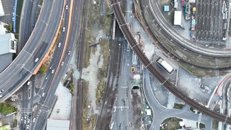 Top-down-shot-from-above-Brisbane-City's-Mayne-Railway-Yard,-as-a-train-passes-through-the-shot,-Busy-car-traffic-on-road-leading-to-ICB-inner-city-bypass