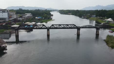 Spectacular-drone-view-of-the-famous-Bridge-on-the-River-Kwai-in-Kanchanaburi,-Thailand-on-an-overcast-day,-revealing-mountains-in-the-distance-through-the-foggy-sky