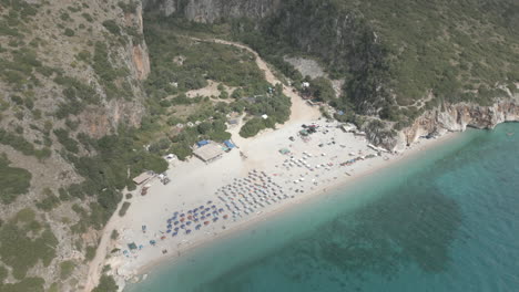 Drone-Disparó-Sobre-La-Playa-De-Gjipe-Albania-Con-Turistas-En-La-Playa-Y-Agua-Azul-Clara-Cerca-De-Las-Montañas-Y-El-Mar-En-Un-Día-Soleado-Y-Brillante-Y-Naturaleza-Verde-Alrededor-Con-Tumbonas-Y-Toallas