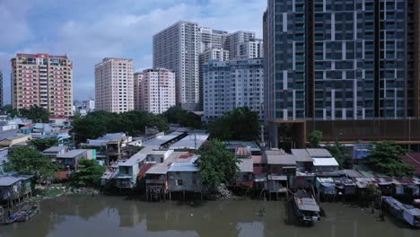 Aerial-view-on-Kenh-Te-canal-Ho-Chi-Minh-City-with-old-iron-and-wood-shacks,-traditional-river-boats-and-ultra-modern-high-rise-buildings-on-sunny-day