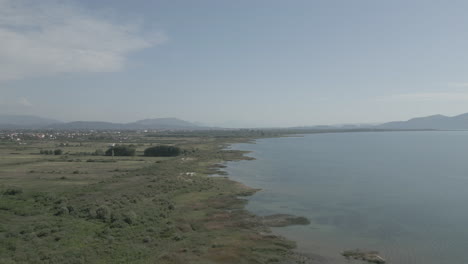 Drone-shot-flying-over-Shkodra-Lake-in-Albania-with-mountains-in-the-background-and-green-plants-grass-nature-underneath-on-a-sunny-day-with-haze-above-the-lake-LOG