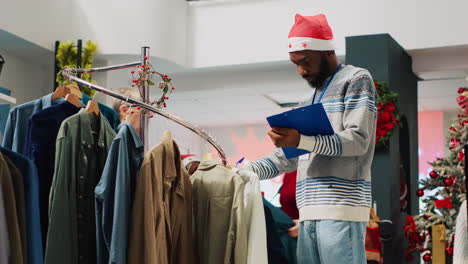 Gerente-Minorista-Navegando-Por-Estantes-De-Ropa-En-Una-Tienda-De-Moda-Decorada-Navideña,-Verificando-Etiquetas-De-Precios-Con-La-Lista-Del-Portapapeles.-Supervisor-Inspeccionando-Blazers-Elegantes,-Buscando-Defectos