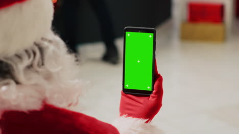 Worker-in-Christmas-ornate-clothing-store-dressed-as-Santa-Claus-holding-green-screen-smartphone,-browsing-through-fashion-shop-ecommerce-website,-inputting-promotional-holiday-sales-online